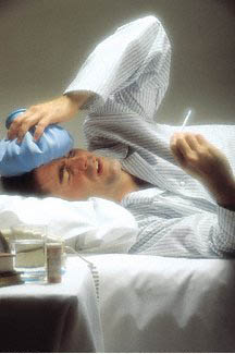 man in pjs lying in bed with icepack on forehead, holding a thermometer and frowning